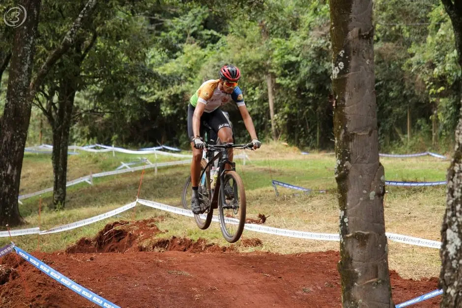 Mountain biker jumps over dirt trail in a forest competition setting.